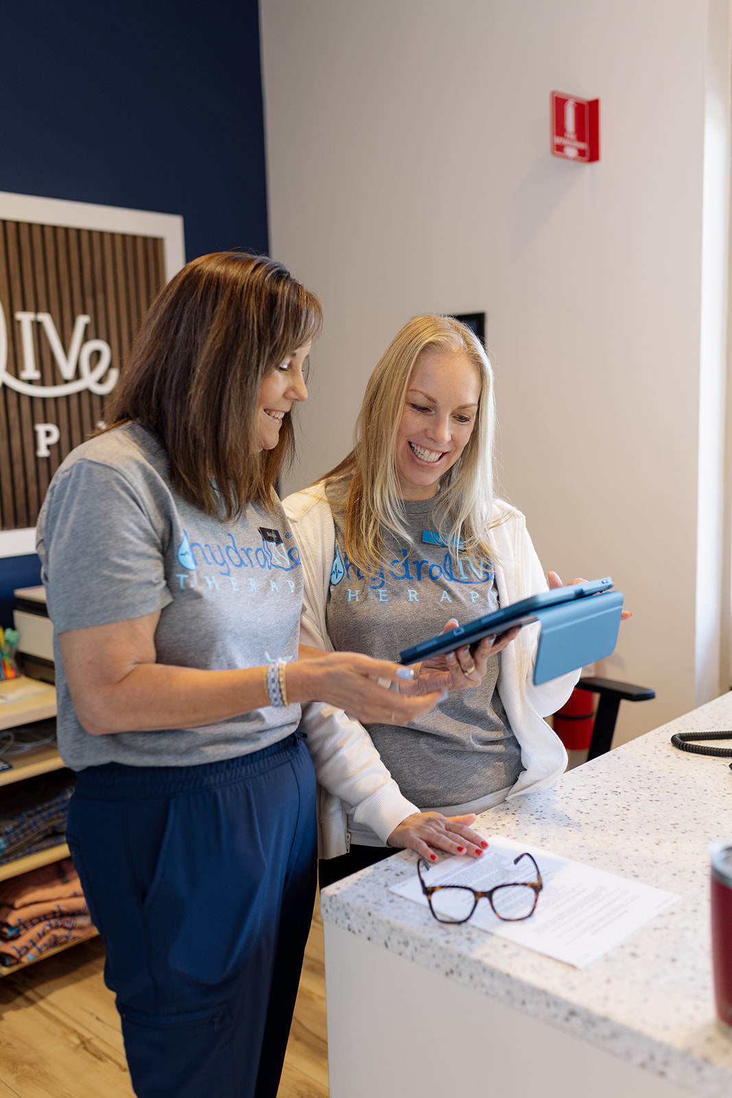 Two Hydralive team members smiling over a tablet at the front desk of the Huntersville clinic