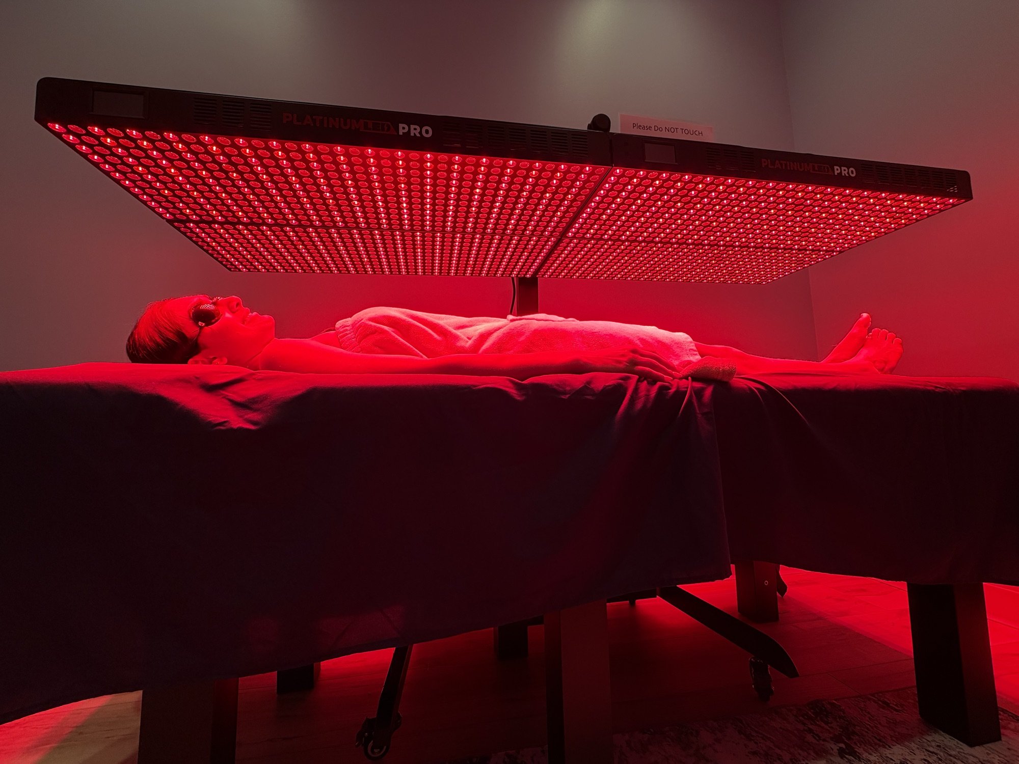 A client lying under the lit PlatinumLED BioMax Pro red light panel array at Hydralive Huntersville, wearing eye protection